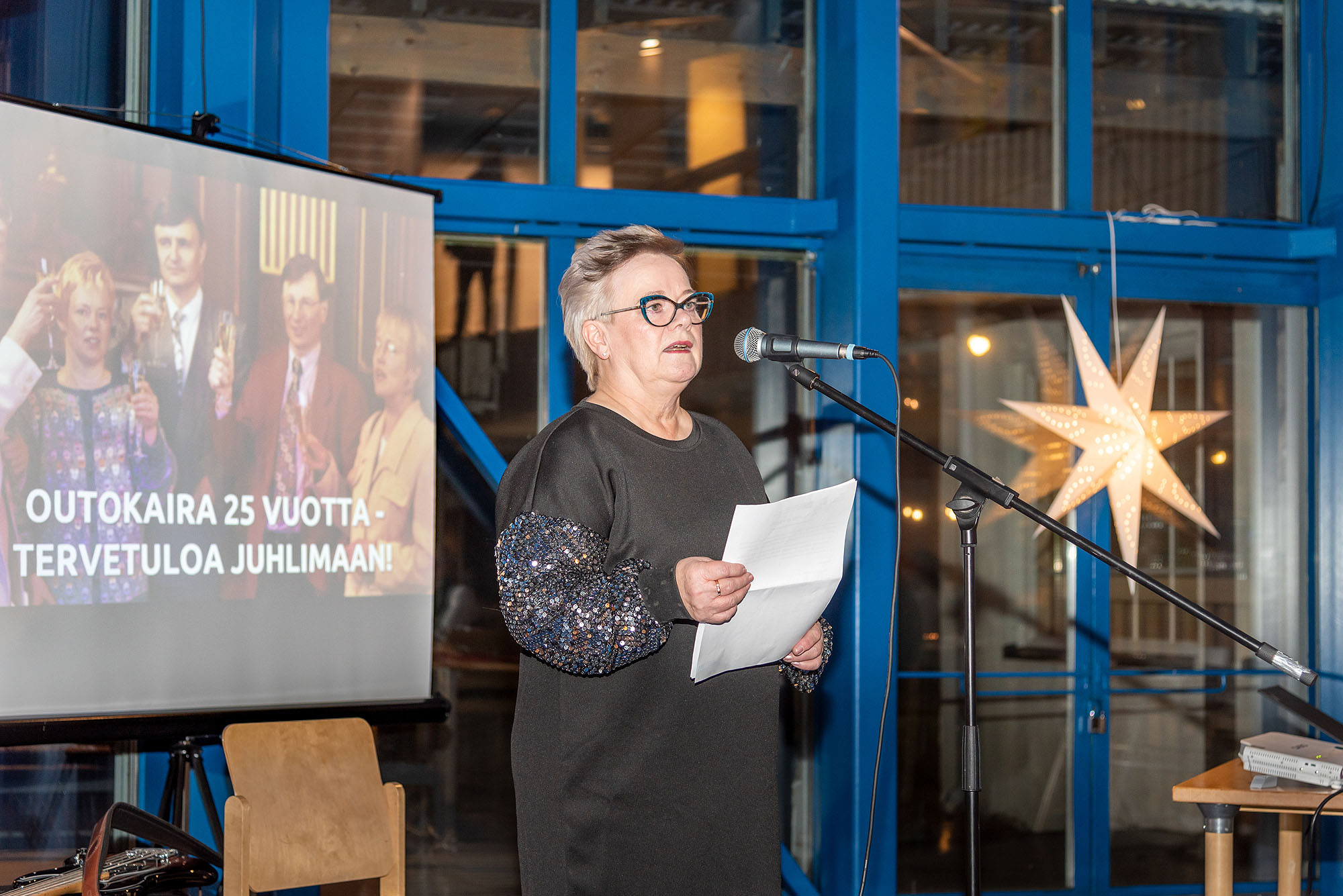 Anne Anttila puhuu juhlassa, taustalla kuva, jossa kilistellään ja lukee Outokaira 25 vuotta - tervetuloa juhlimaan!